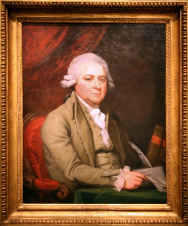 John Adams, posing for a portrait while wearing a tan jacket and white undergarment. This American Revolutionary era painting depicts Adams as a lawyer and middle-aged patriot. As a man of law and integrity, the painting is encased a gold frame, exemplifying his stature in society. Additionally, Adams is seen donning a white wig; a practiced custom of British Law.