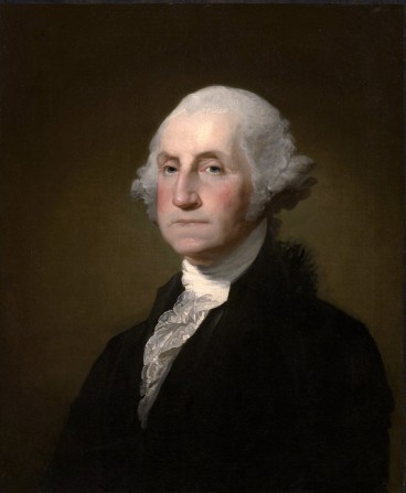 George Washington, posing for a portrait, is wearing a black jacket with a white undergarment. This painting is from later in his life, which can be inferred due to his grey hair and aging facial appearance.